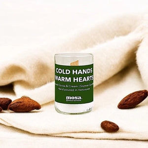 Glass candle with 'Cold Hands Warm Hearts' text on a soft fabric background with almonds.