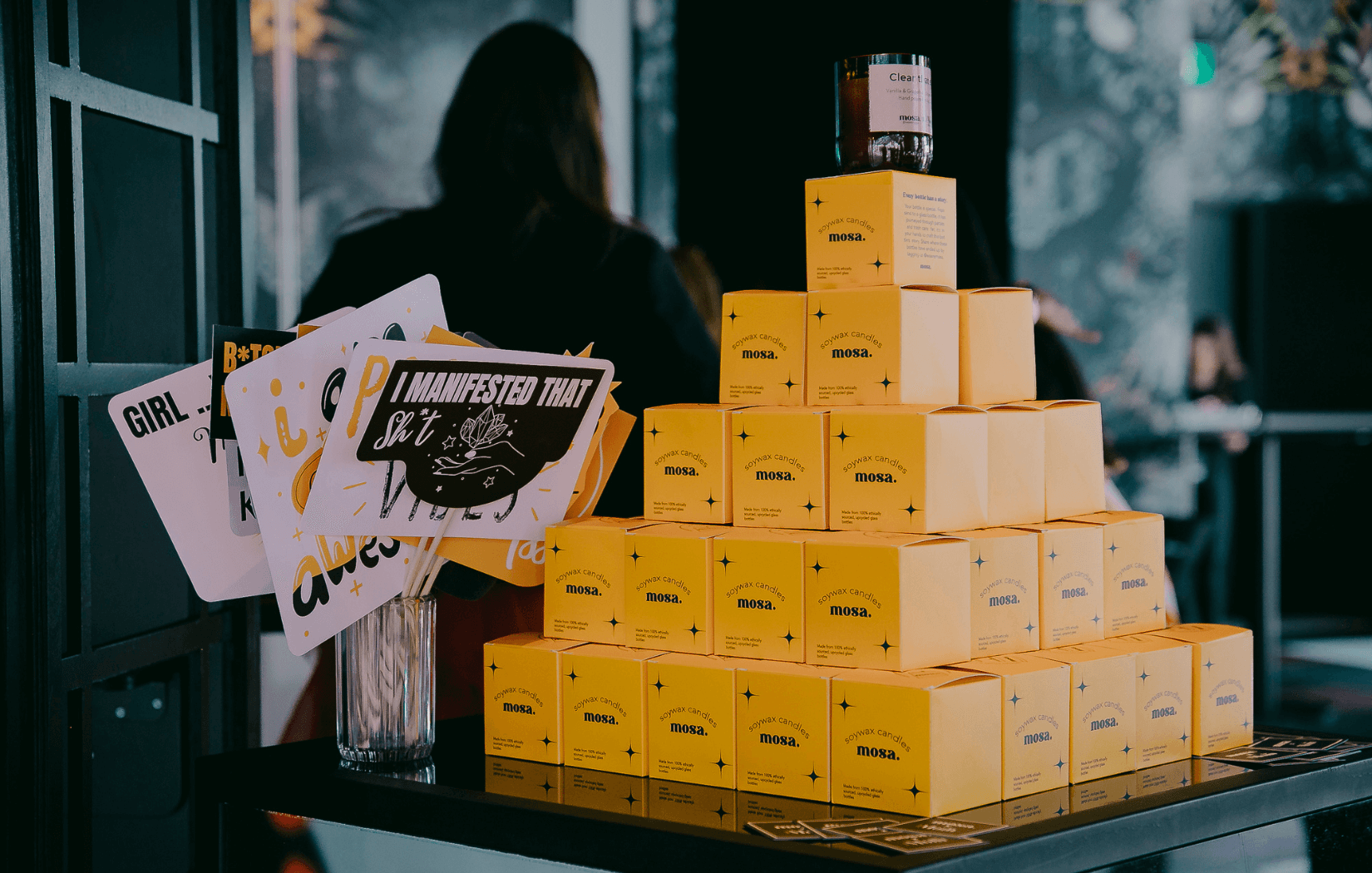 A display pyramid of bright yellow Mosa soy wax candle boxes arranged neatly on a black counter, topped with a single open candle jar. Beside it, a clear glass holds fun, illustrated affirmation signs with phrases like “I manifested that s**t” and “Girl Power,” set in a stylish indoor event space.