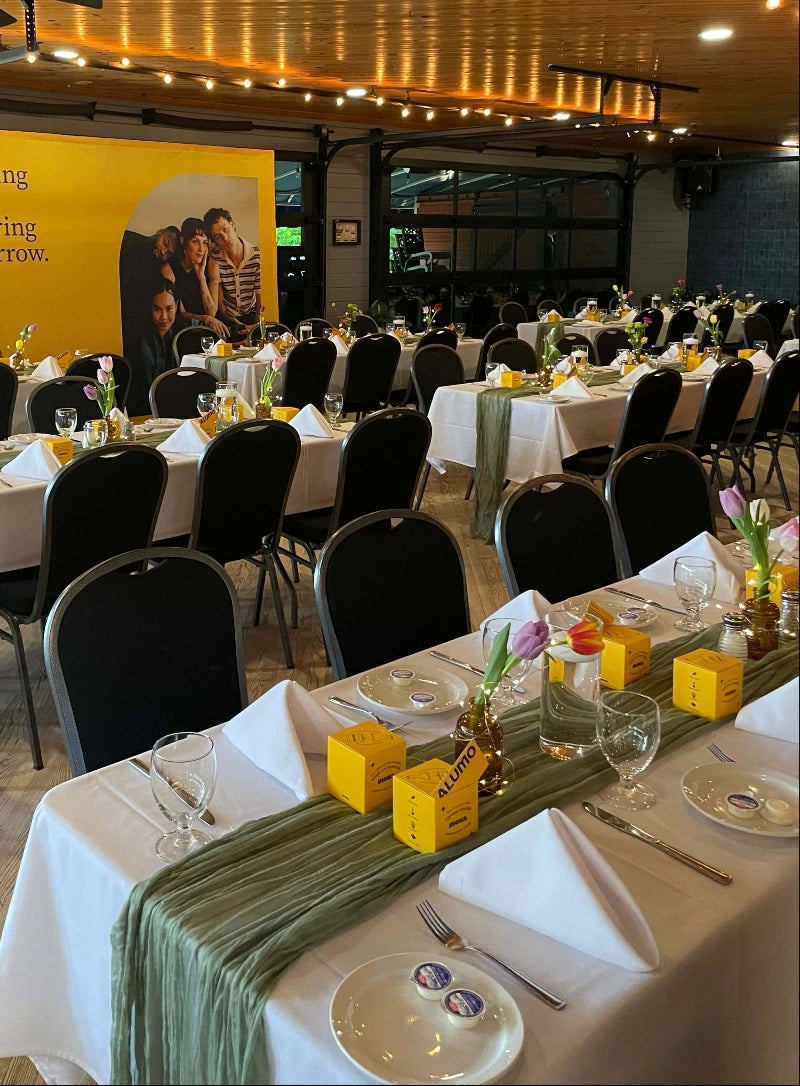 Dining hall with tables set for a meal, featuring yellow Mosa sustainable soy-wax candle boxes and green table runners.