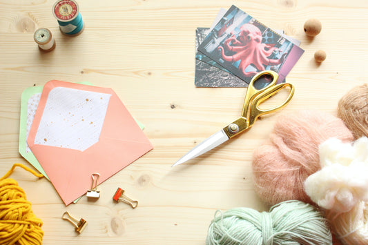Various crafting supplies laid out on the surface of a wooden table, including postcards, scissors, sewing thread, envelopes, binder clips, and differently coloured spools of yarn. 