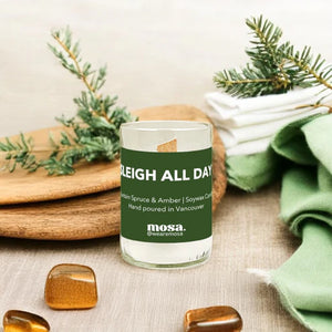 Candle with 'Sleigh All Day' label on a surface with greenery and stones
