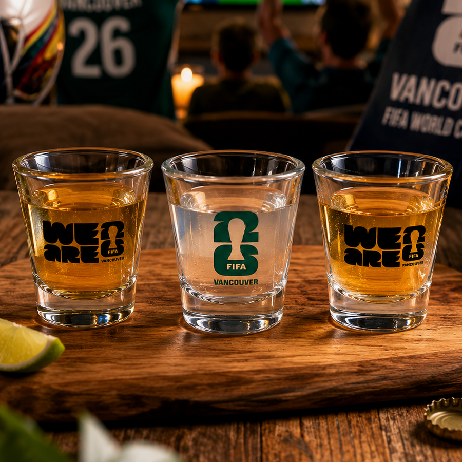 Three shot glasses with FIFA branding on a wooden board, with a soccer ball and World Cup paraphernalia in the background.