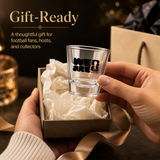Empty shot glass with 'WE ARE FIFA' text held by a hand, gift-wrapped in brown paper with gold ribbon, against a dark background.