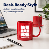 Red mug with 'WE ARE 2026 VANCOUVER' text on a desk with a laptop and pen.