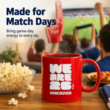 Red mug with 'WE ARE 26 VANCOUVER' text on a table with popcorn and people watching sports on TV.