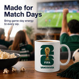 White mug with FIFA Vancouver logo on a table with popcorn and a soccer ball, people watching TV in the background.